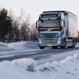 A blue truck going down a snowy road lined with trees.