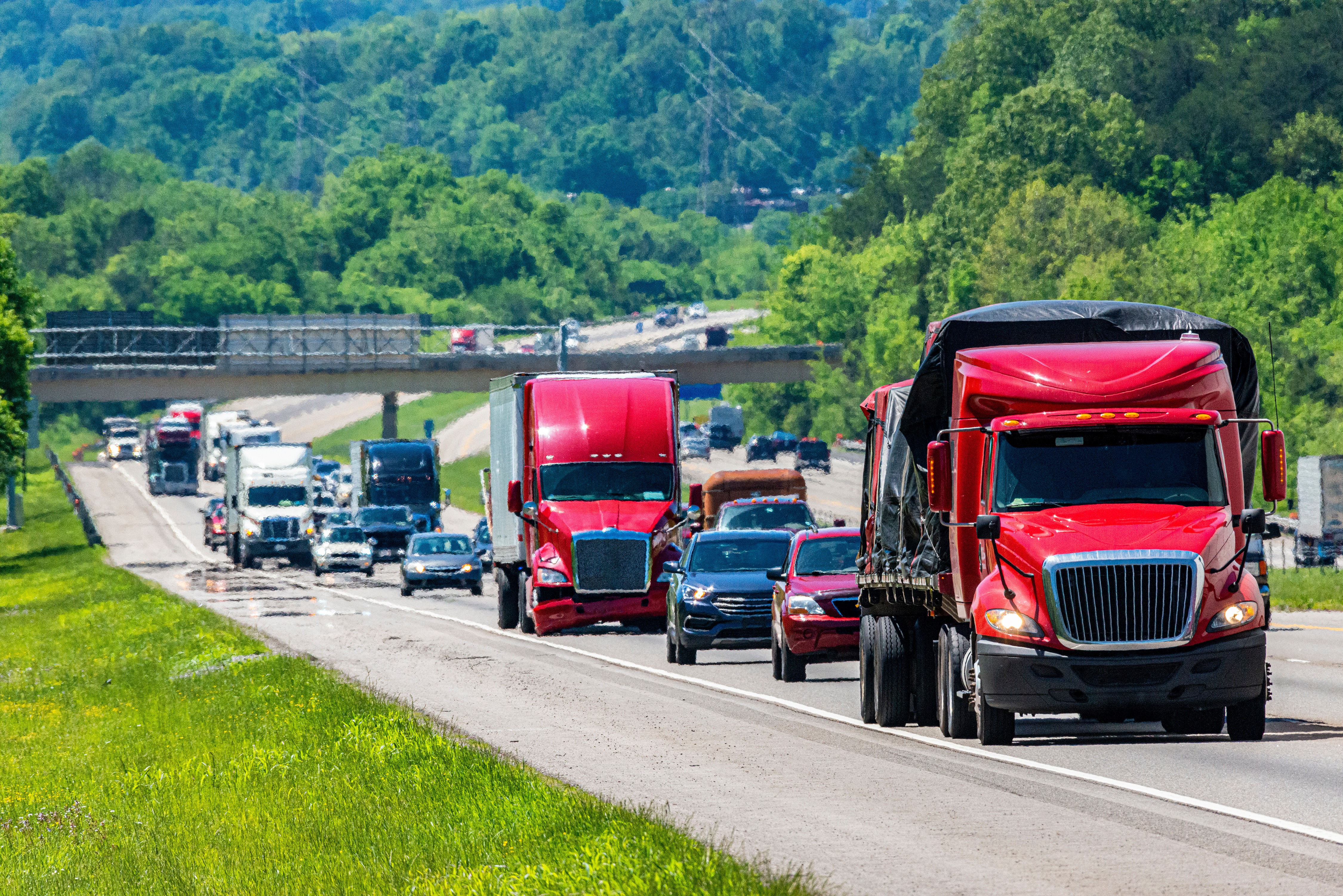 Red semi-trucks and passenger vehicles driving on a highway through green forested landscape