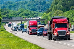 Red semi-trucks and passenger vehicles driving on a highway through green forested landscape