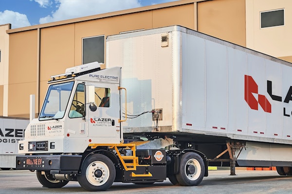 An Orange EV yard tractor hauling a Lazer Logistics trailer at a loading dock.