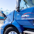 A blue NFI-owned Volvo VNR Electric parked at a shipping port.