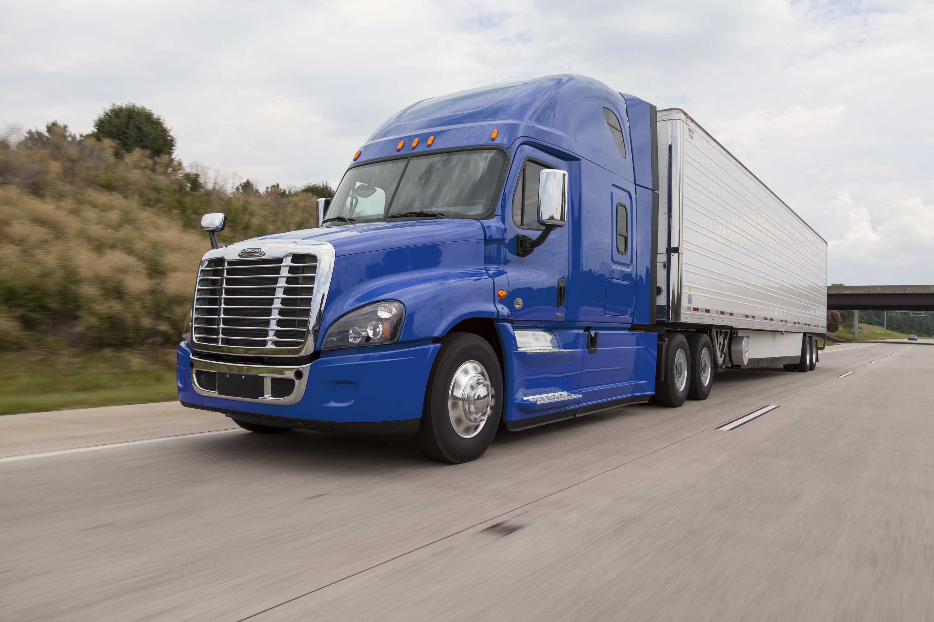 A blue Freightliner Class 8 semi truck hauling a silver trailer driving on a highway.