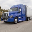 A blue Freightliner Class 8 semi truck hauling a silver trailer driving on a highway.