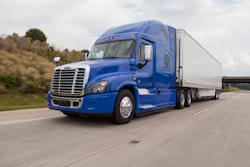 A blue Freightliner Class 8 semi truck hauling a silver trailer driving on a highway.
