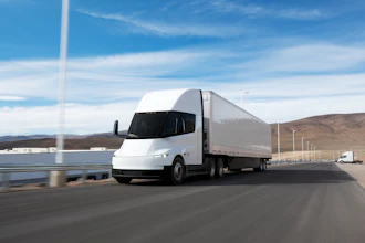 A white Tesla Semi hauling a trailer driving along a coastal road.