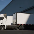 A lineup of Einride battery-electric semis with trailers attached parked at a warehouse loading dock.