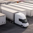 A Tesla Semi with a trailer is parked in a logistics yard alongside other trucks.